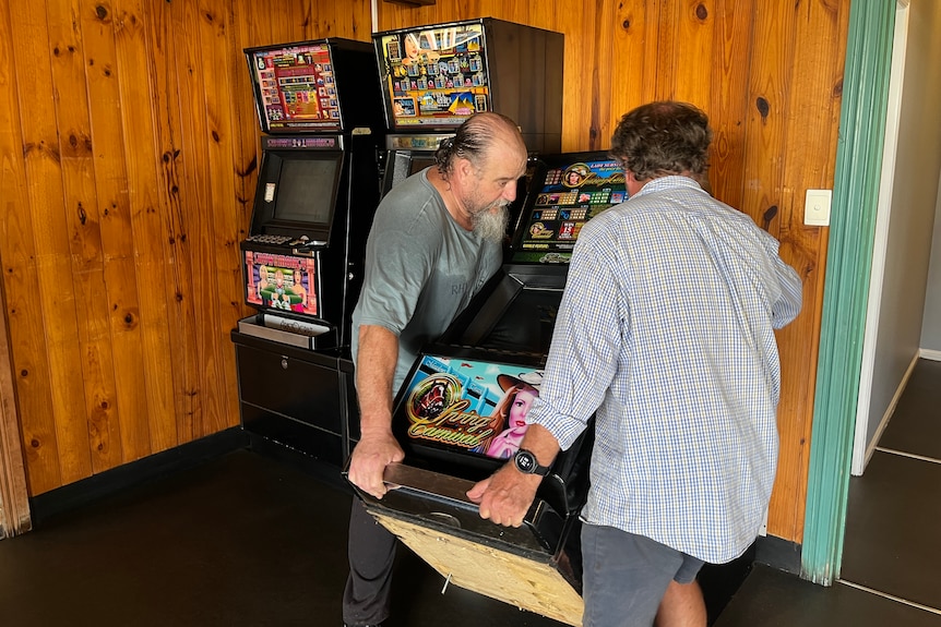 two men lift a gaming machine onto a trolley in a room with brown wooden walls