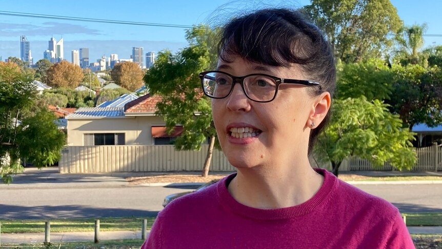 Gabrielle is standing on a Perth street. It’s sunny and she had her dark hair tied back and is wearing black-rimmed glasses.