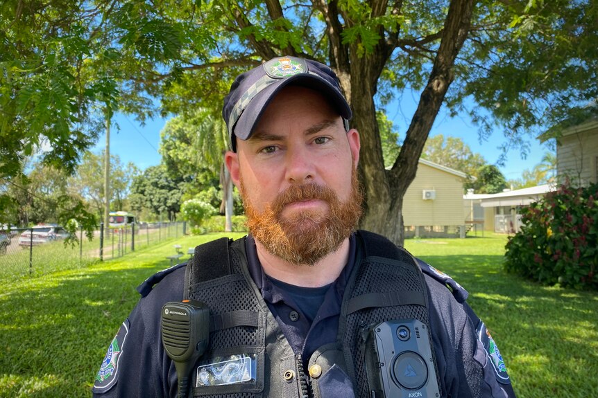 A bearded police officer stands under a tree in front of a school.