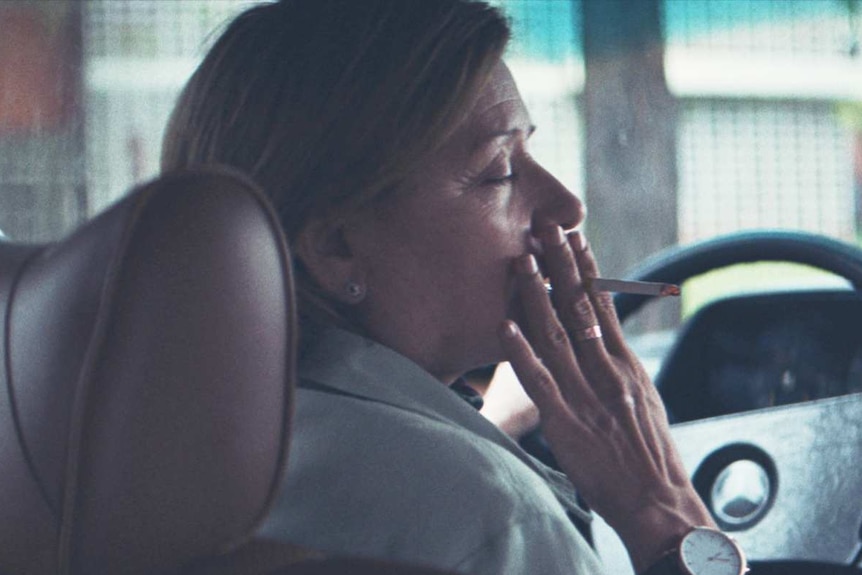 Two women sit in the driver and passenger seat of a car, the woman in the passenger seat holds a cigarette to the driver's lips.