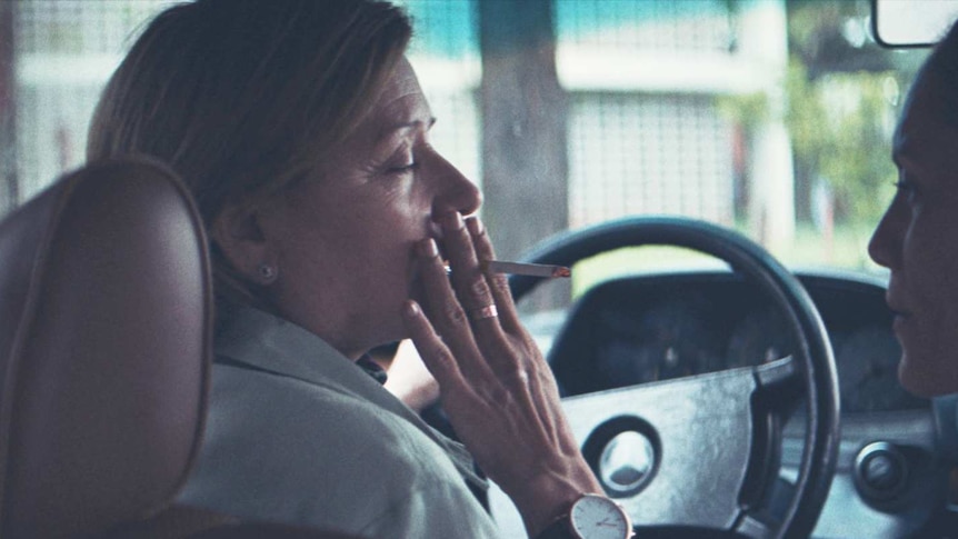 Two women sit in the driver and passenger seat of a car, the woman in the passenger seat holds a cigarette to the driver's lips.