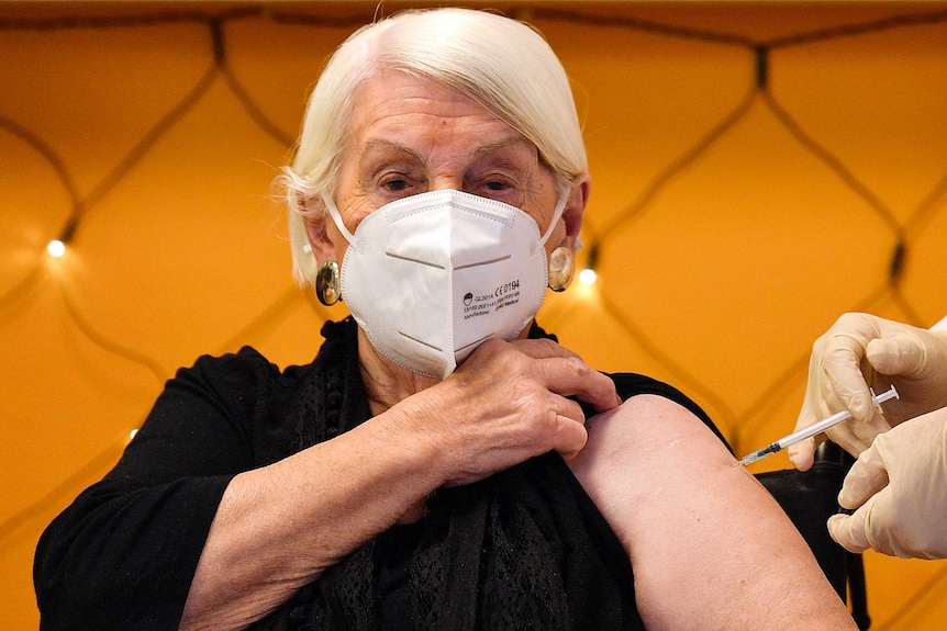 An elderly woman with white hair and wearing a mask pulls up her black shirt while someone administers a needle to her arm.