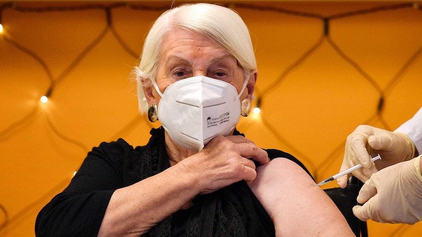 An elderly woman with white hair and wearing a mask pulls up her black shirt while someone administers a needle to her arm.