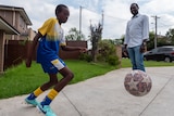 A boy kicks a soccer ball in the driveway while a man in a shirt watches on. 