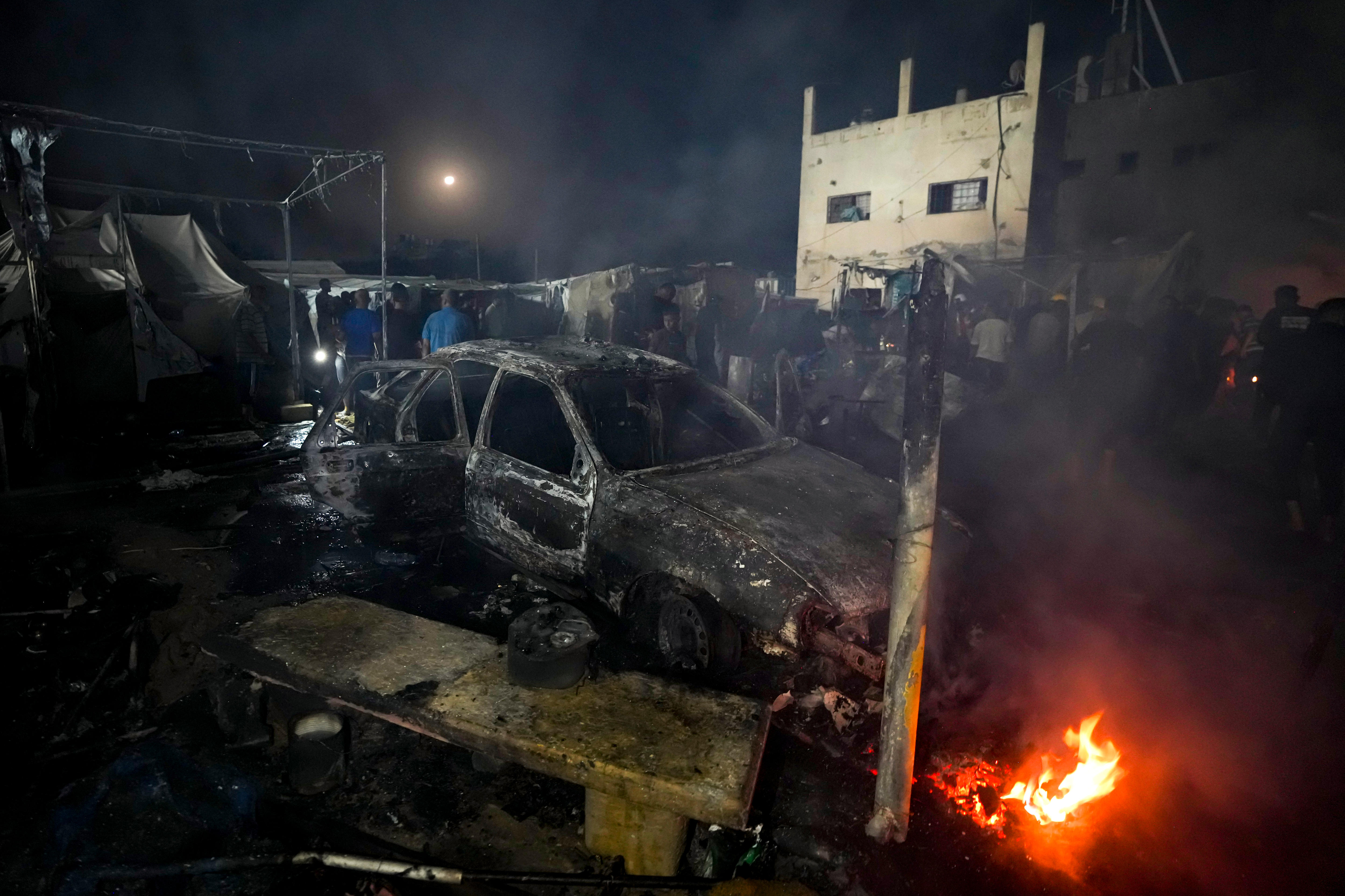 A burn-out car stands in the centre of a heap of wreckage including burned tents