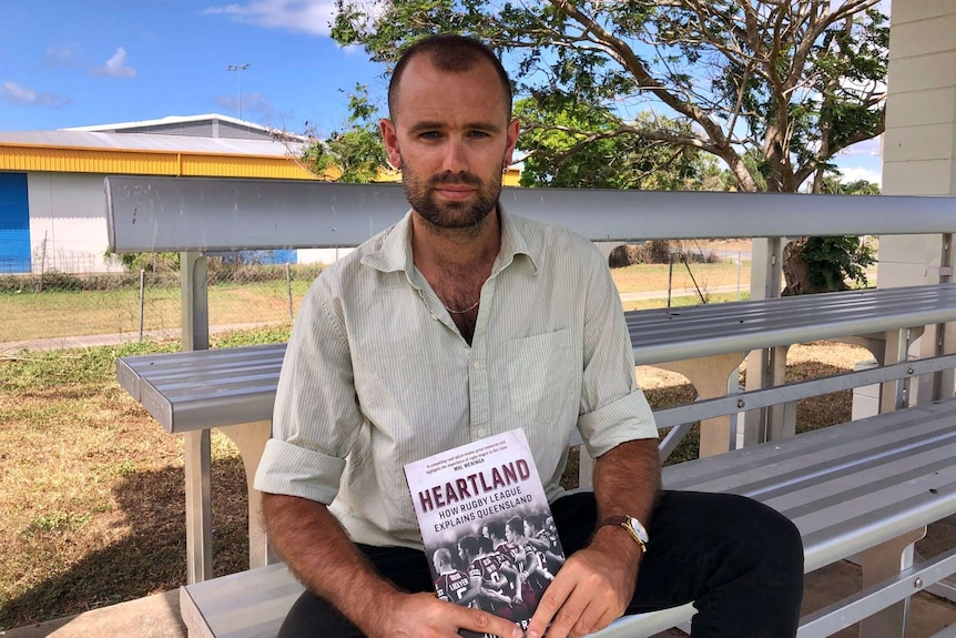 Man sitting on a metal bench holding a book entitled Heartland: How Rugby League Explains Queensland