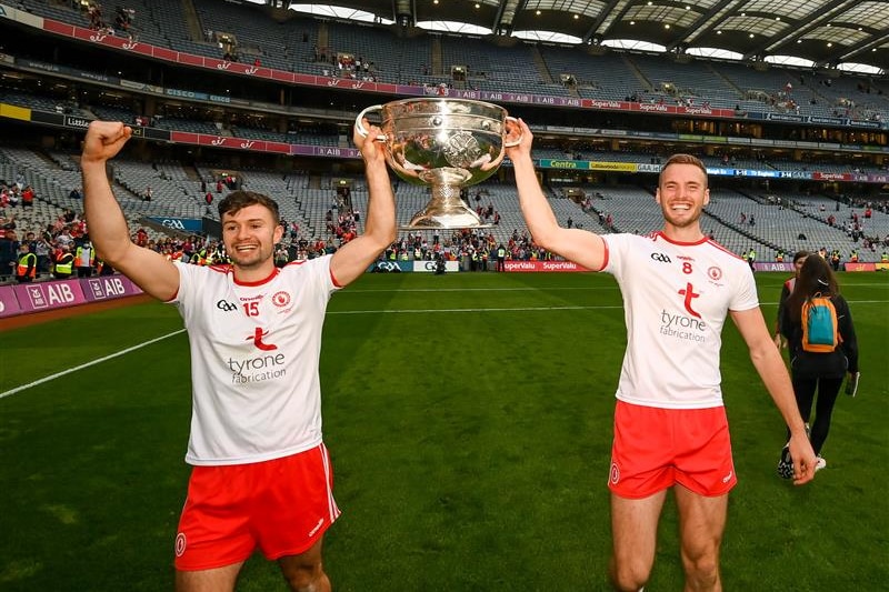 Two Tyrone players walk on the ground of Croke Park after the All-Ireland final, holding the trophy between them.