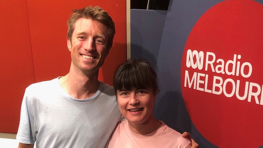 A man in a grey tee shirt has his arm around a woman in a pink tee shirt in a radio studio. They are both smiling for the camer