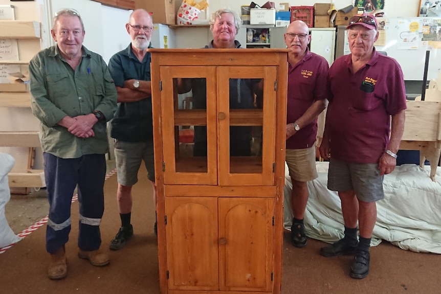 Five men stand smiling around a large, wooden cupboard.