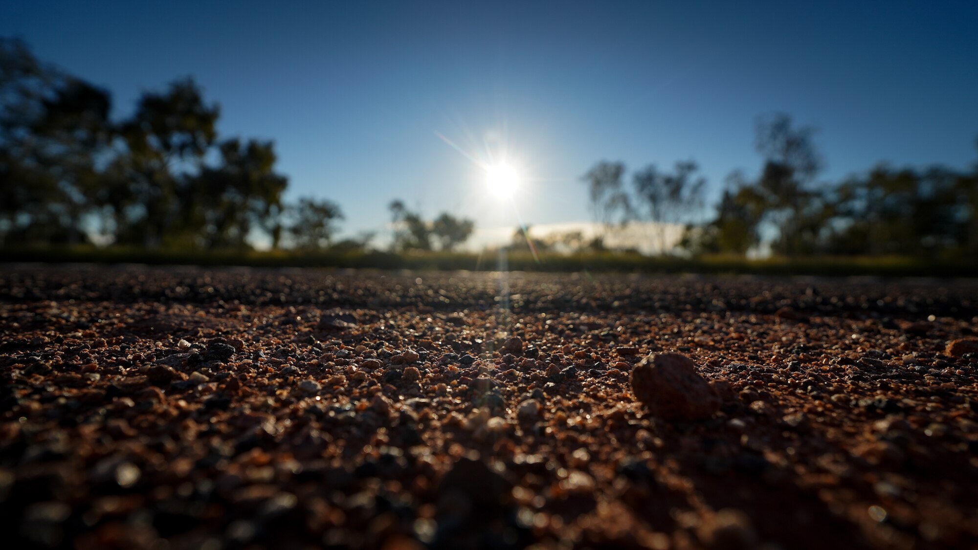 A gravel road close up, brown pebbles in view as the sunlight pours down on the bitumen.