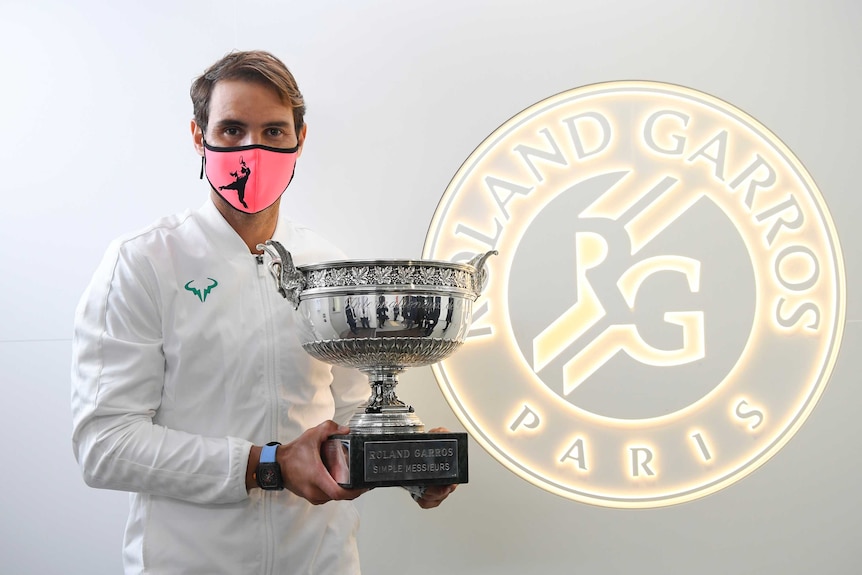 A tennis player wearing a pink mask holds the French Open trophy next to a Roland Garros logo.