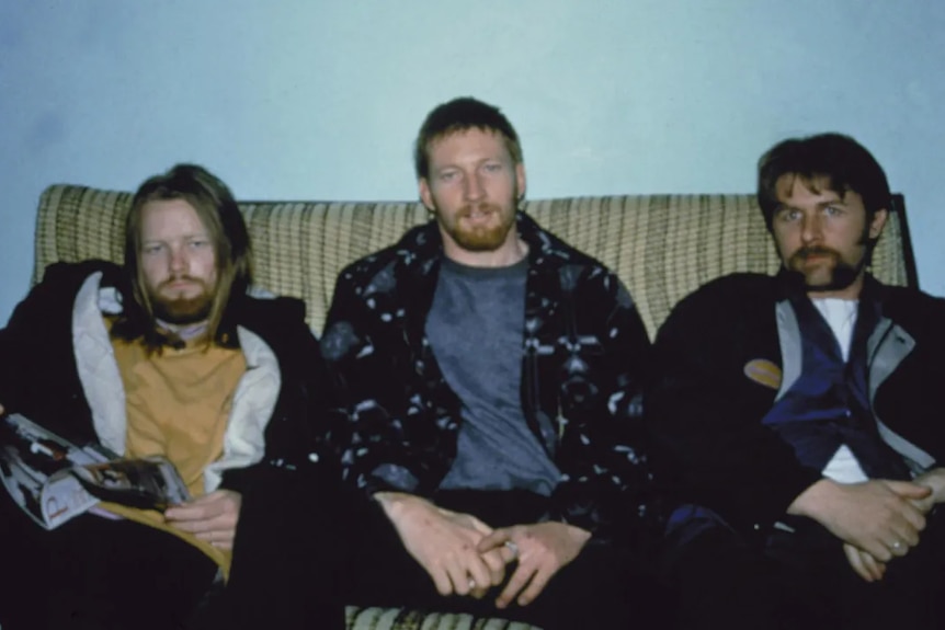 Three young men look into the camera. They're seated on a lumpy couch, legs spread wide. They all have facial hair.