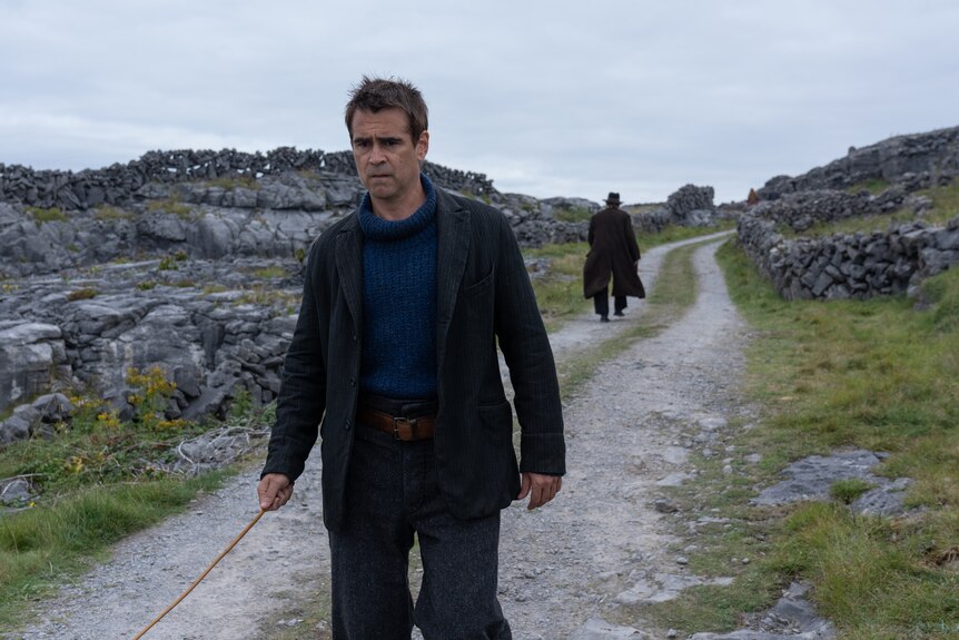 A middle-aged man in 20s-style dress is walking, looking upset, through the Irish landscape, an older man walking away from him.