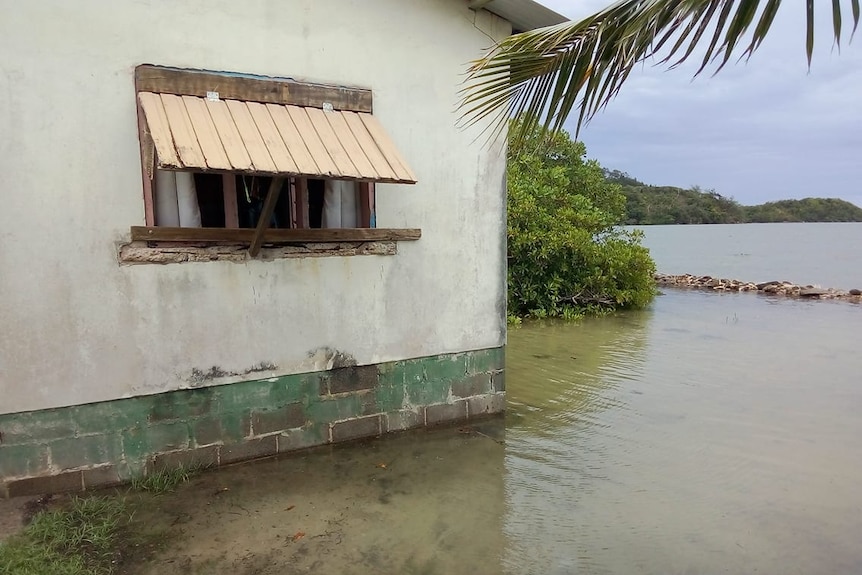 The corner of a house with a palm tree and the sea surrounding the lower part of the house.