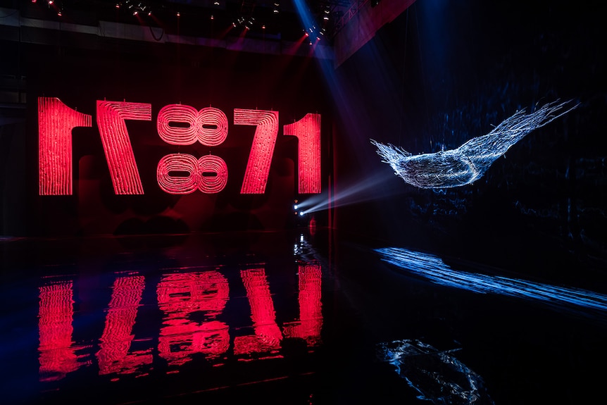 A red sculpture reading 1788 and nest made of branches are illuminated and suspended in dark exhibition space.