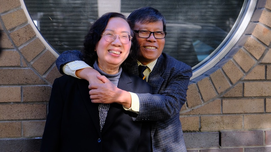 A Vietnamese Australian man smiles with his hands embracing his smiling wife. 