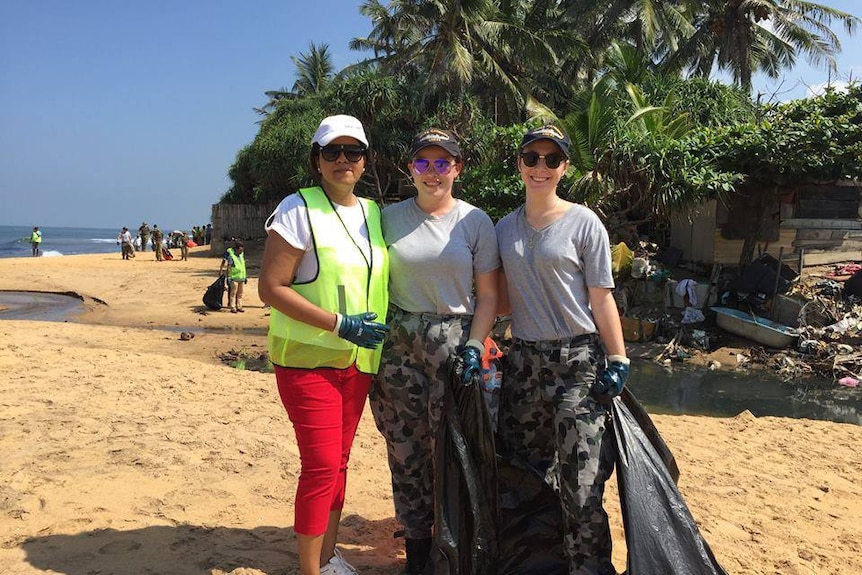 Manik in a bright yellow vest, sunglasses and cap on a beach next to two women in army camouflage