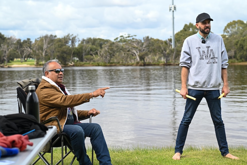 A man sits in a chair pointing towards a man standing holding tapping sticks, on the banks of a calm river. 