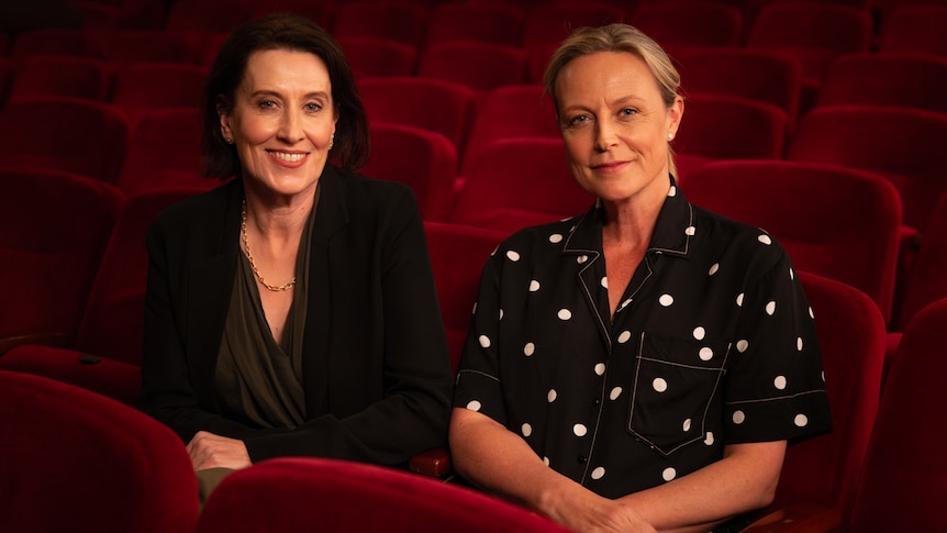 Virginia Trioli and Marta Dusseldorp, two middle-aged women, sit together smiling in a theatre, on a bank of red chairs.