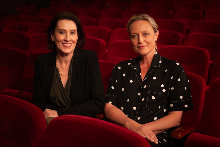 Virginia Trioli and Marta Dusseldorp, two middle-aged women, sit together smiling in a theatre, on a bank of red chairs.