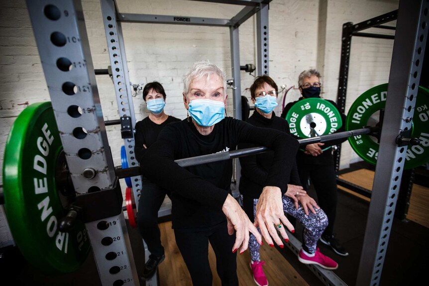 Four women in facemasks to protect against coronavirus stand behind a weight machine