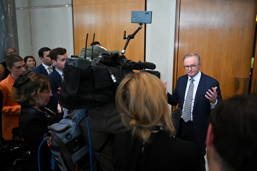 A man at the centre of a media scrum, gesticulates and answers questions