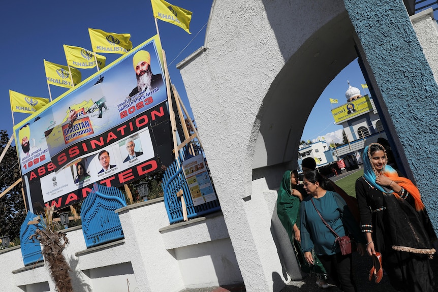 A  group of women stand near a billboard erected on a wall outside a temple. 