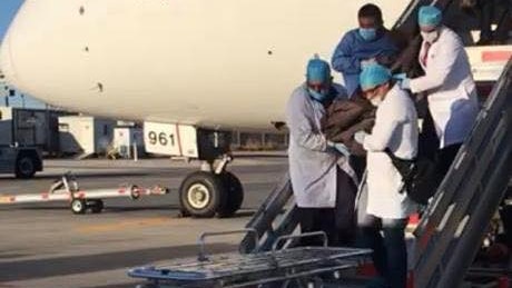 People wearing medical scrubs carry a body down the stairs of a plane to a waiting stretcher.