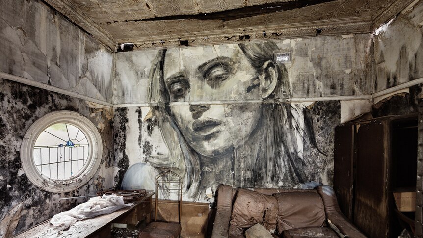 A painting of a woman looks down from a wall with peeling paint and wallpaper over a room of disused furniture