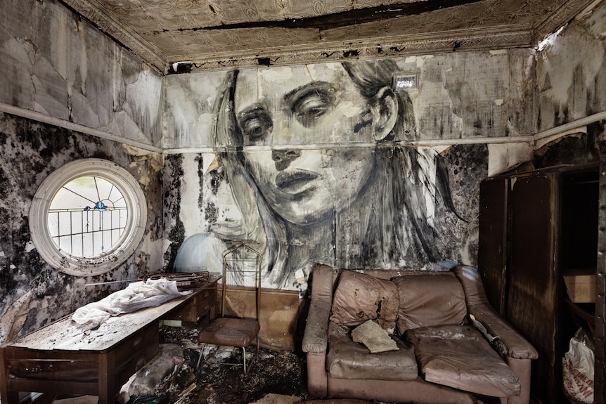 A painting of a woman looks down from a wall with peeling paint and wallpaper over a room of disused furniture