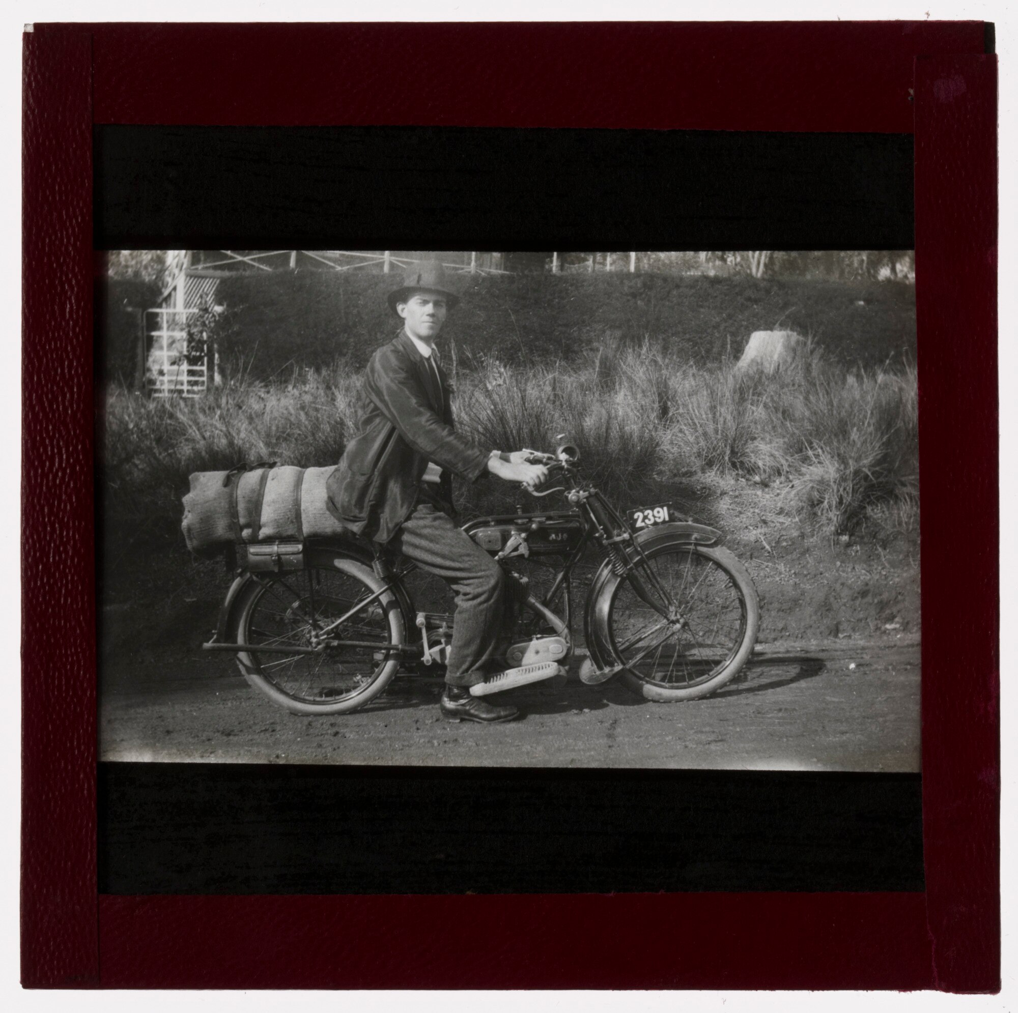 Nevill Westwood sits on a bike with a large bag on the back. He wears a jacket and hat. The photograph is black and white.