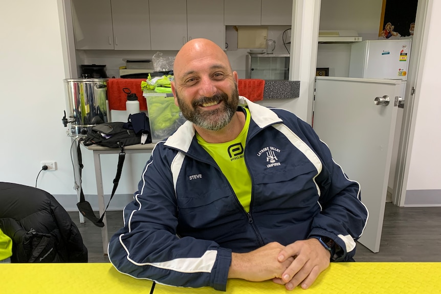 A smiling bald man sitting behind a table in a sports clubroom