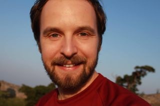 A close up photo of a bearded man with brown hair smiling at the camera while wearing a red t-shirt and standing outside.