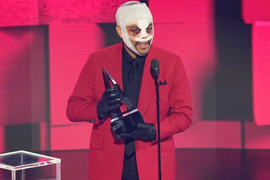 With bandages all over his head and wearing a red suit, The Weeknd holds a trophy and speaks into a microphone