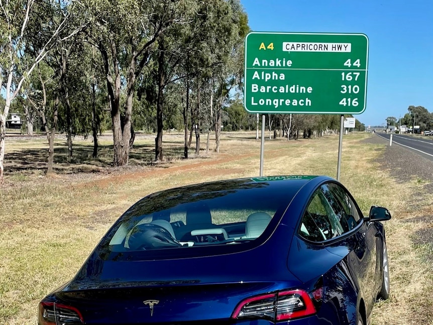 a navy blue tesla sedan parked in front of a sign which reads "anakie, alpha, barcaldine, longreach"