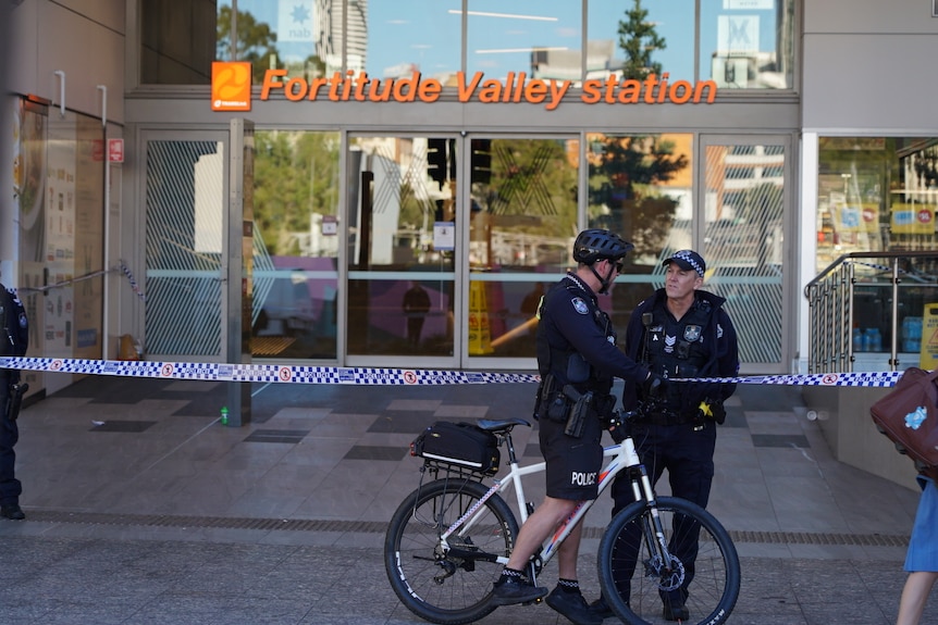 Two police men, one on a bicycle, stand outside the Fortitude Valley police station with police tape.