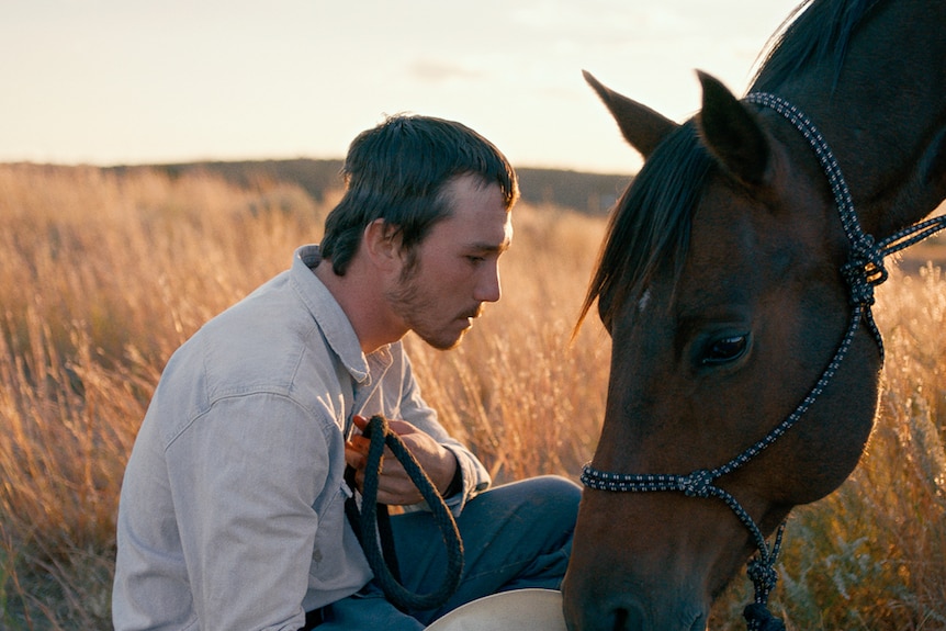 A man crouching in field with his head close to a horse's downward-leaning head.