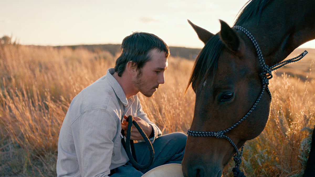 A man crouching in field with his head close to a horse's downward-leaning head.