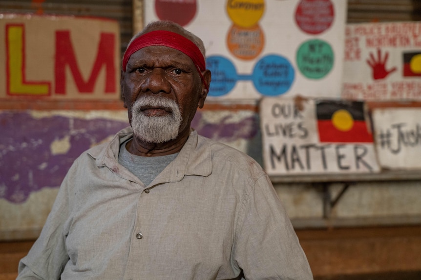 An Indigenous man with a white beard and red headband looks at the camera