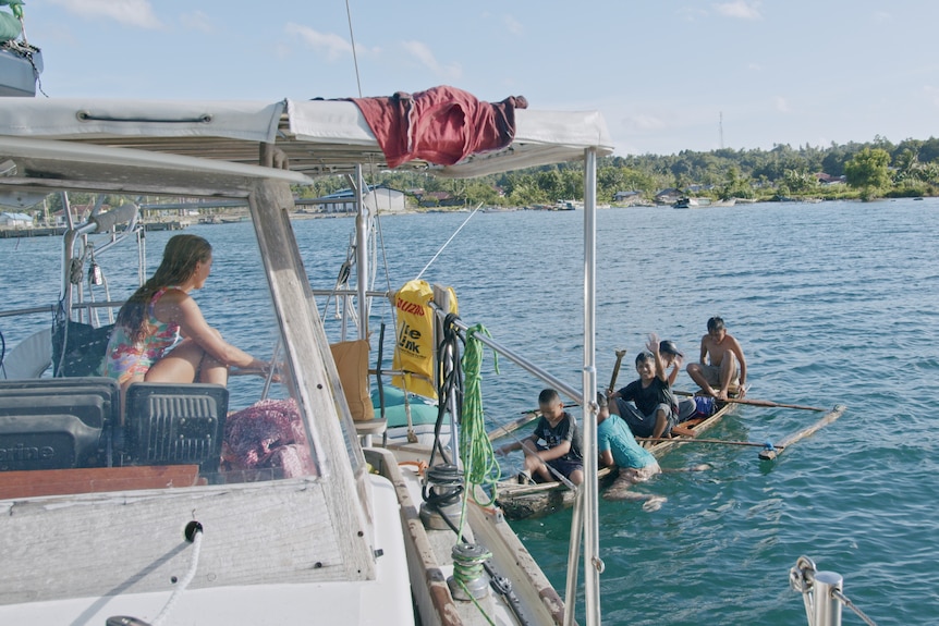 A group of boys in a small boat near a larger boat with a woman on board.