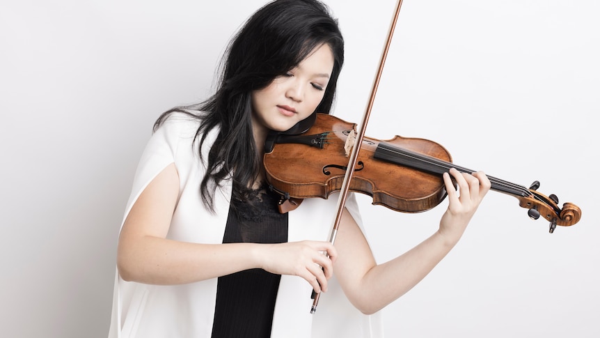 Violinist Emily Sun wearing a white jacket and playing her violin with her eyes closed.