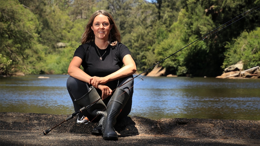 Erin Lake sits next to the Kangaroo Valley River holding a fishing rod.