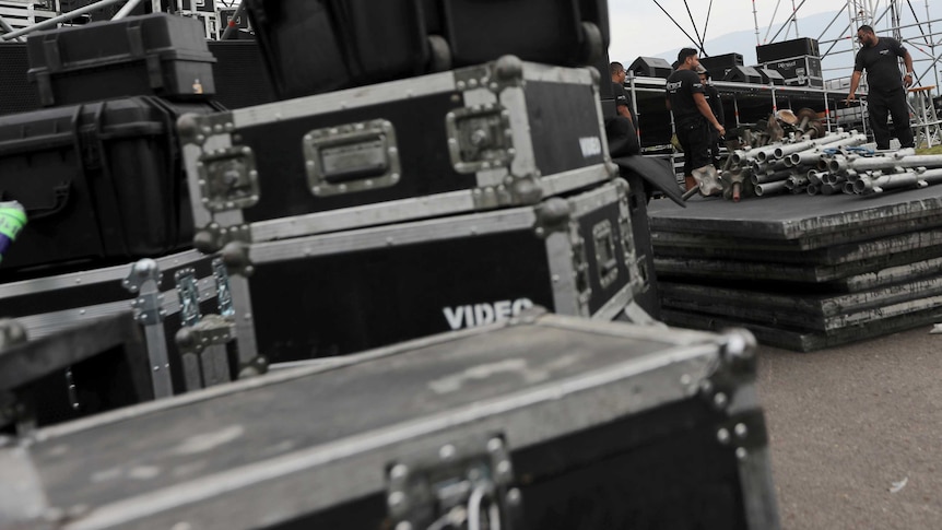 Crates are stacked on top of each other before a concert. Men in the background set up.