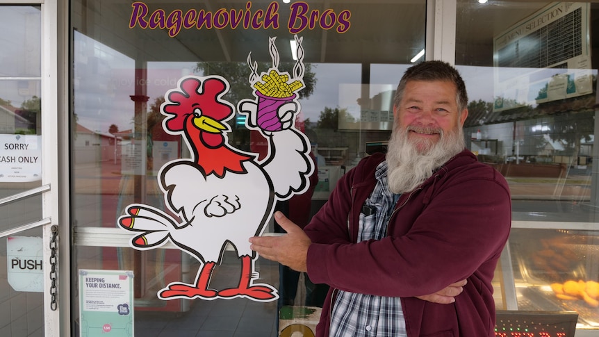 A bearded man smiles outside the window of a chicken shop.