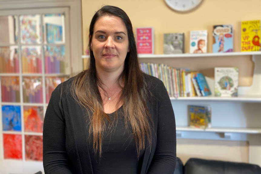 A woman with long hair wearing a black top stands in front of children's books on shelves