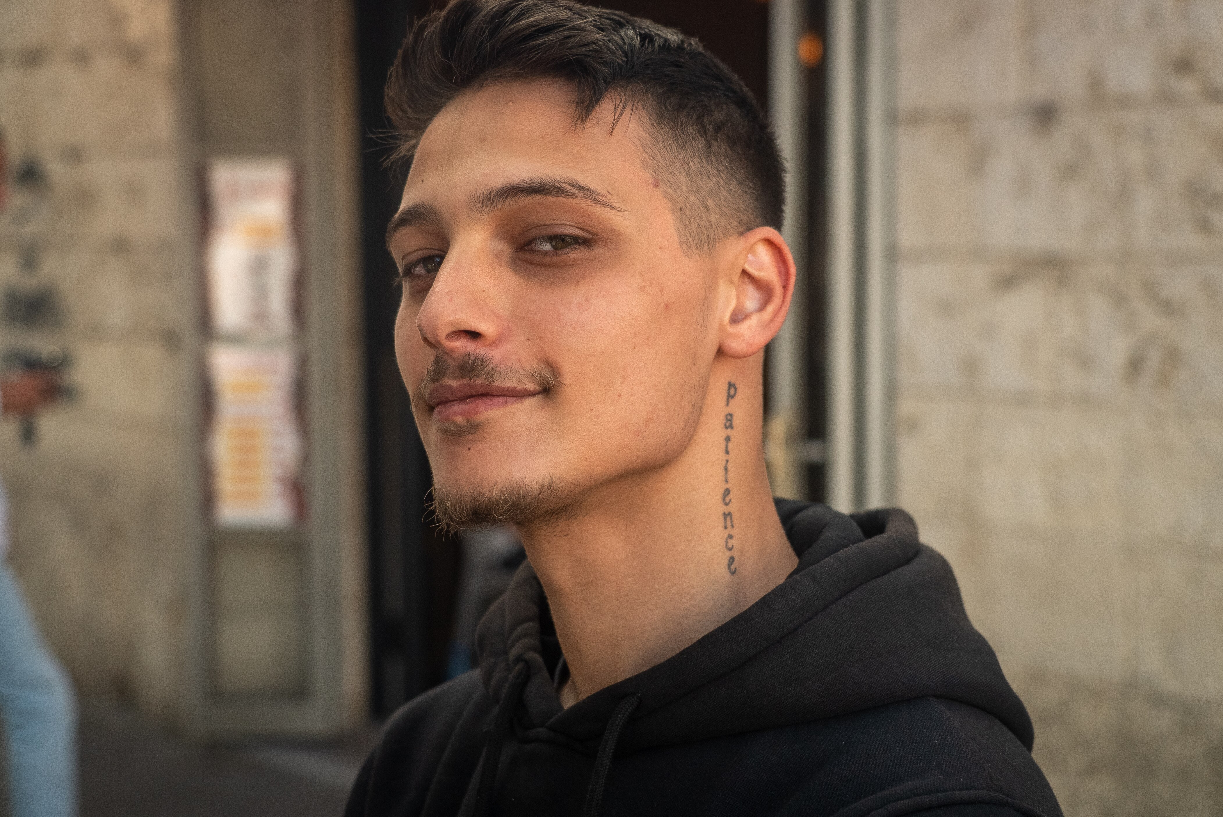 A man gives a small smile and tilts his chin slightly upward. 'Patience' is tattooed down his neck