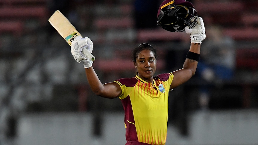 A female cricketer raises her bat in one hand and helmet in the other during a match and smiles