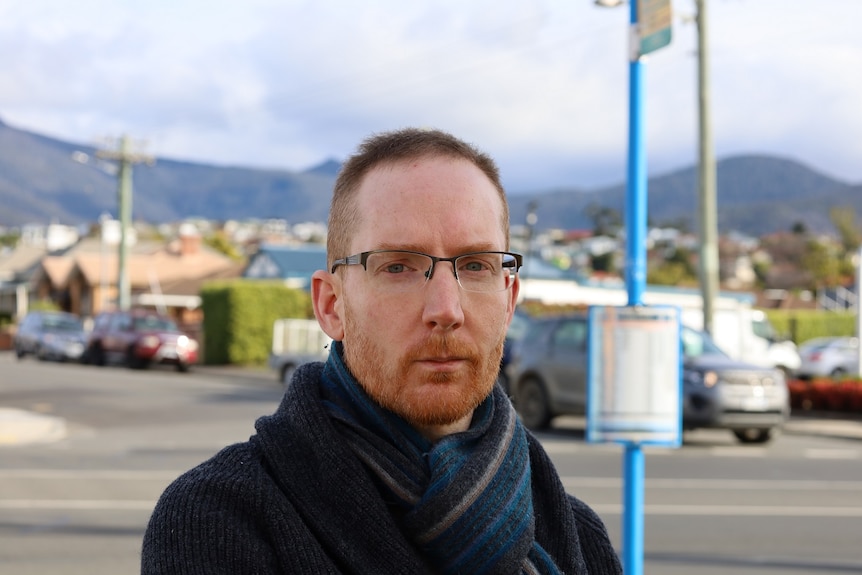 A man at a bus stop looks at the camera, with an out of focus bus timetable, traffic and houses in the background