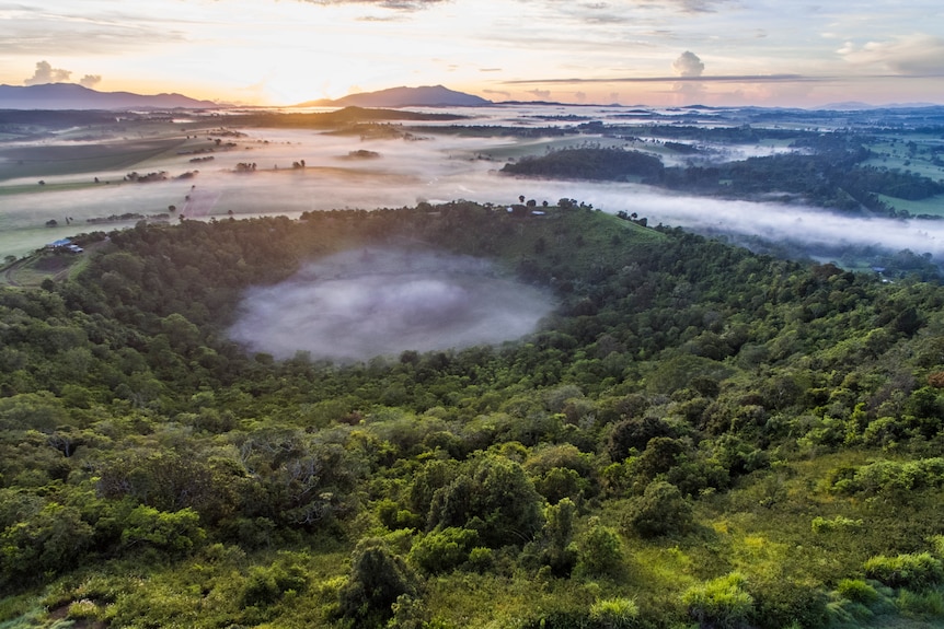 An aerial shot of a crater a top a large green mountain 