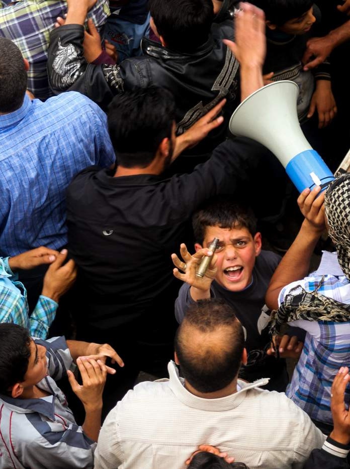 You look down at a crowd of men and young boys huddled, while a young boy looks up aggressively while holding a bullet.